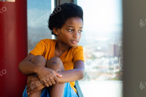 Preview: Thoughtful biracial schoolboy wearing yellow tshirt and looking through window in classroom