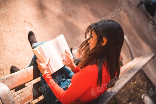 Preview: Young Caucasian brunette in a red shirt sitting on a bench in the park reading a book