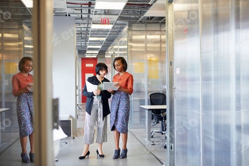 Preview: Two businesswomen in office corridor, looking at digital tablet