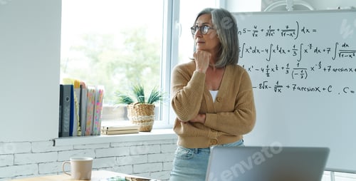 Preview: Thoughtful senior woman holding hand on chin while standing near the whiteboard at classroom