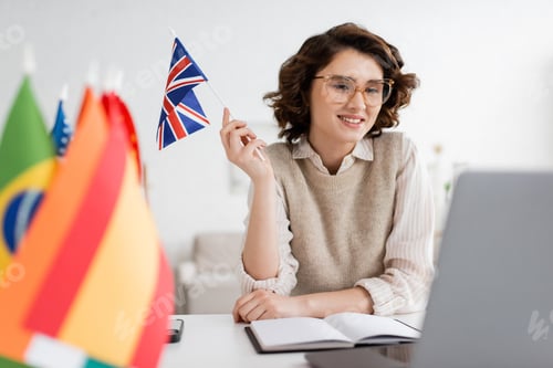 Preview: smiling language teacher in eyeglasses holding flag of United Kingdom near notebook and blurred