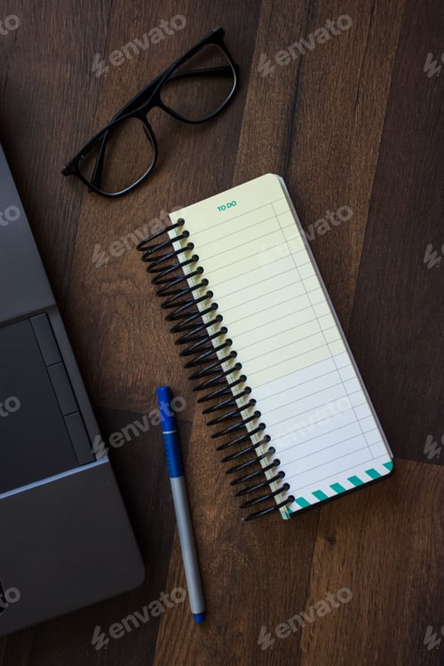 Preview: Top view of a wooden desk with a notebook, a pen, and eyeglasses next to a laptop computer.
