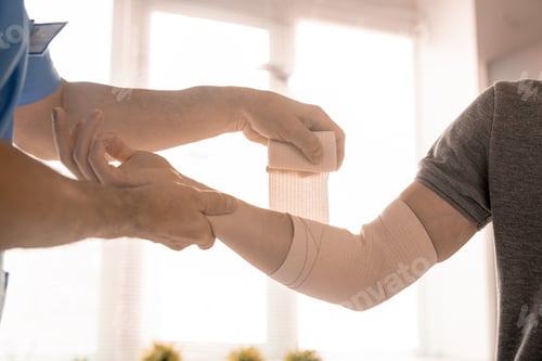 Preview: Hands of clinician wrapping elbow of right arm of sick young man with bandage