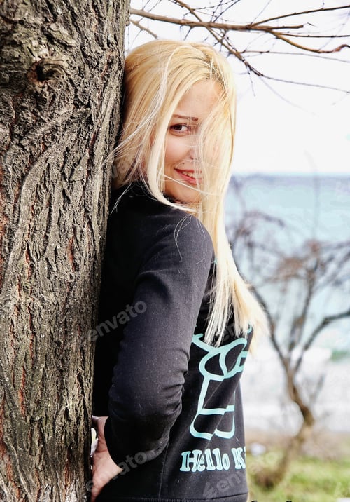 Preview: A young beautiful woman is standing near the tree and smiling