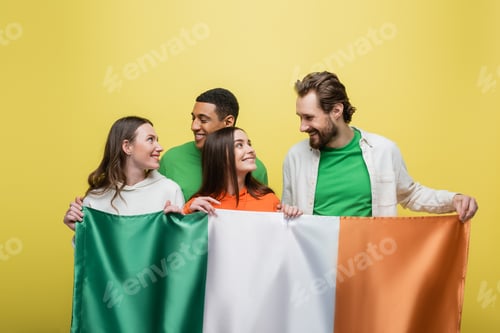 Preview: Interracial friends looking at each other while holding Irish flag isolated on yellow