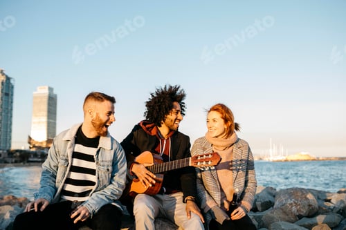 Preview: Three happy friends with guitar sitting outdoors at the coast sunset