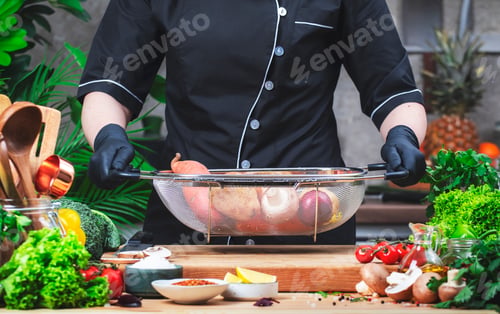 Preview: Cook holds mesh colander with vegetables. Cozy kitchen with wooden table, kitchenware
