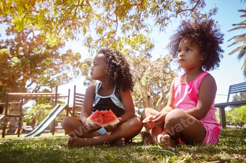 Preview: Two young sisters, sitting on grass, eating watermelon