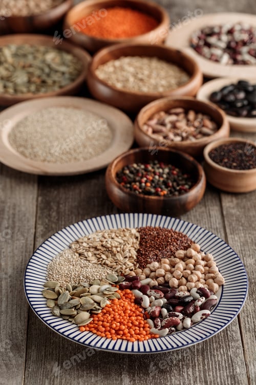 Preview: striped plate with lentil, beans, chickpea, cereals and pumpkin seeds near wooden bowls on table