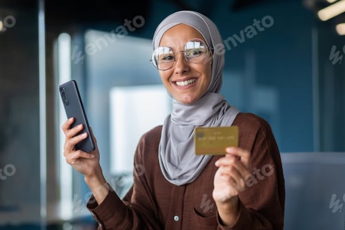 Preview: Portrait of smiling young Muslim woman in hijab sitting in office. Holds a phone in his hands