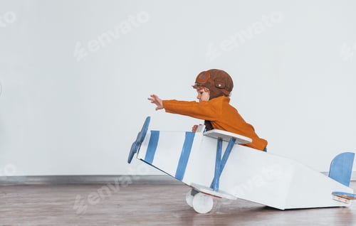 Preview: Little boy in retro pilot uniform having fun with toy plane indoors