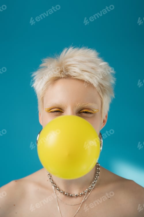 Preview: blonde albino woman with yellow eye liner blowing bubble gum on blue background