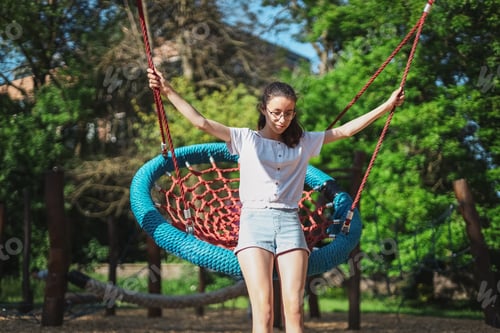 Preview: Portrait of a beautiful caucasian teenage girl sitting down on a round rope swing in the park