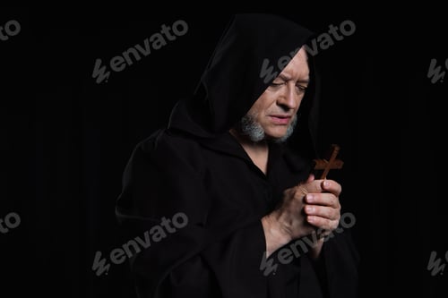 Preview: senior monk in hood looking at wooden cross while praying isolated on black