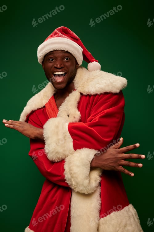 Preview: overjoyed african american man in santa claus costume gesturing and looking at camera on green