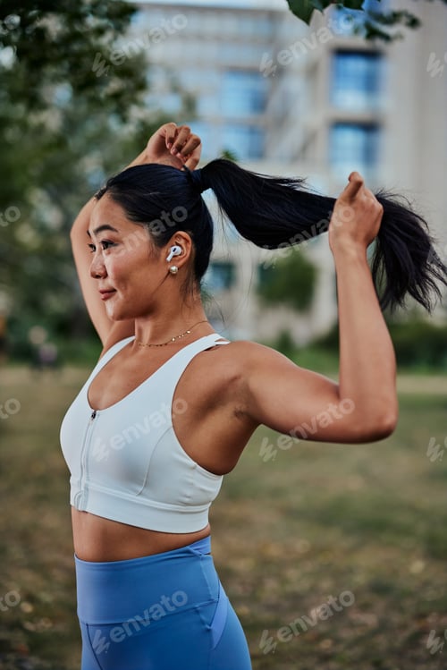 Preview: Attractive asian sportswoman standing in park and tying hair and preparing for workout in park