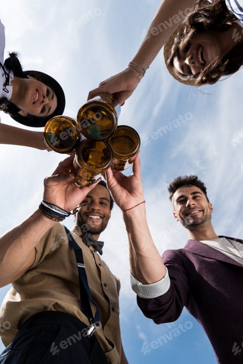 Preview: Low angle view of happy young friends clinking beer bottles outdoors