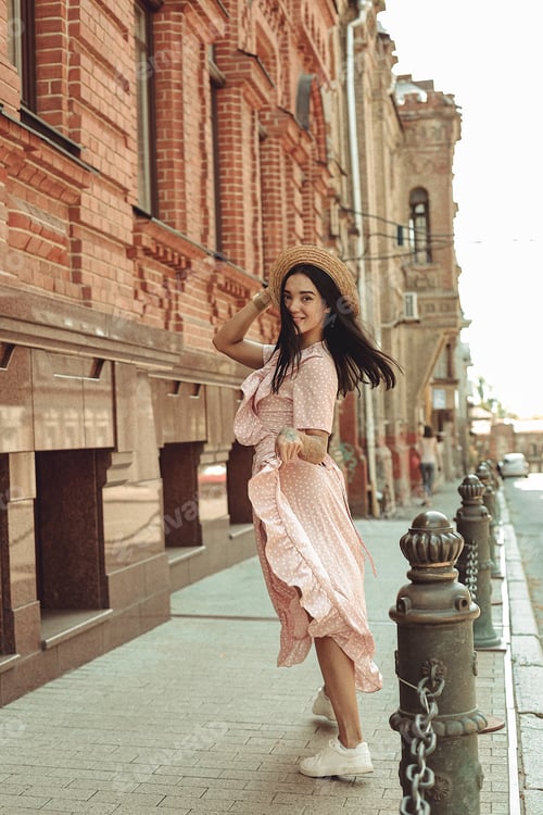 Preview: A young brunette in a soft pink dress with a hat walks around the city