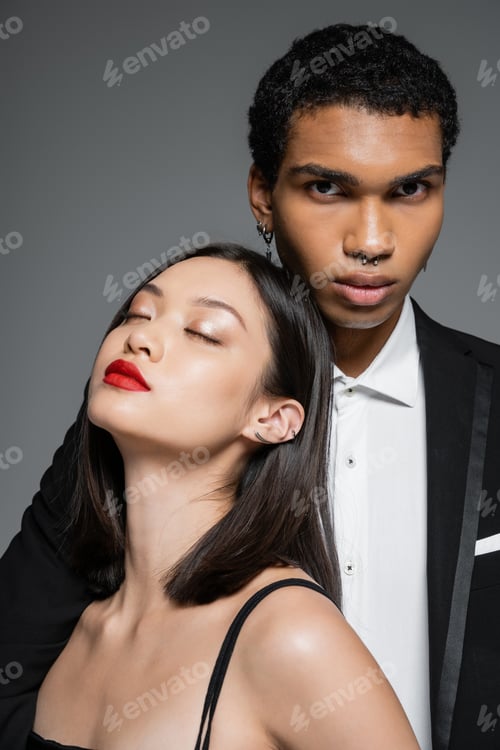 Preview: stylish african american man looking at camera near sensual asian woman with red lips and closed