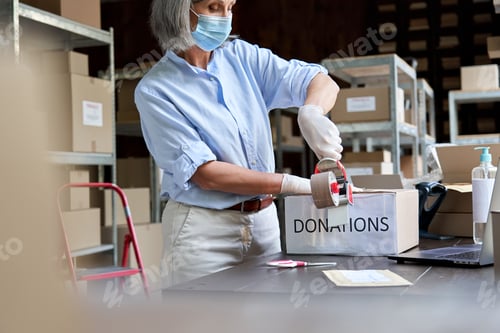 Preview: Mature female warehouse worker wearing mask packing donations box.