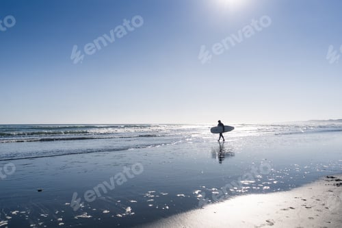 Preview: Backlit man walking along the beach with a surfboard under his arm