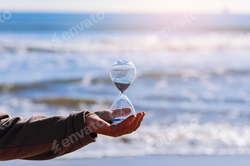 Preview: Time is running fast. Hourglass with blue sand inside in mature woman's hand symbolize the brevity