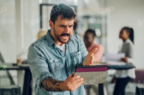 Preview: Angry business man using tablet in the office with his team working in the back