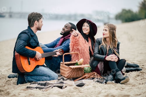 Preview: Happy young friends sitting on sandy beach and enjoying guitar at alfresco picnic