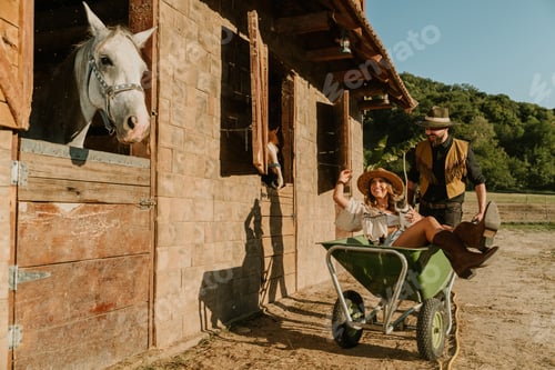 Preview: Cheerful cowboy is driving cowgirl in wheelbarrow at horse ranch.