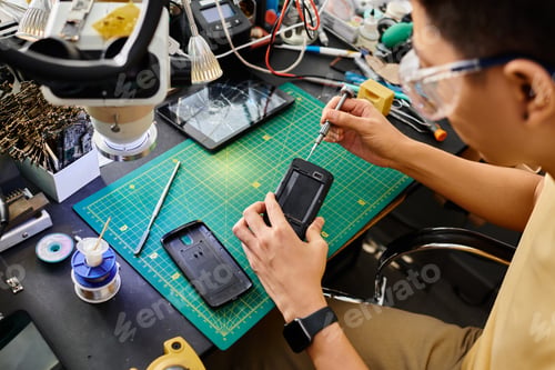 Preview: technician in goggles with screwdriver repairing mobile phone at workplace in private workshop