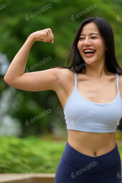 Preview: Happy fitness woman showing her biceps while smiling in a green park