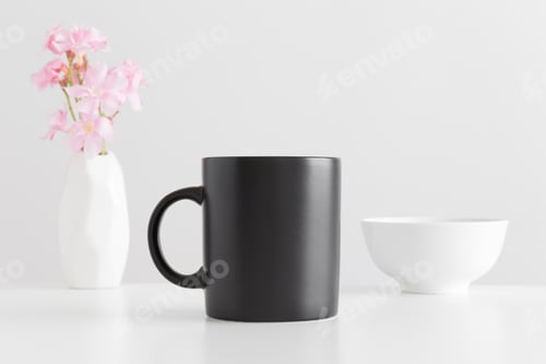 Preview: Black mug mockup with a bouquet of a pink oleander in a vase and a bowl on a white table.