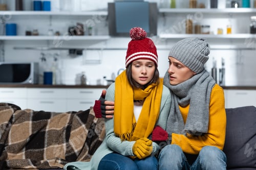 Preview: young man in warm hat and scarf hugging freezing girlfriend while sitting in cold kitchen
