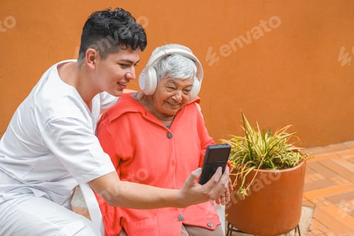 Preview: Young caregiver teaching elderly woman to use smartphone and headphone