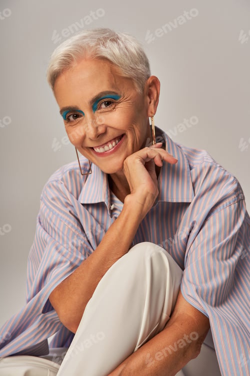 Preview: cheerful senior model with bold makeup posing in blue striped shirt and looking at camera on grey