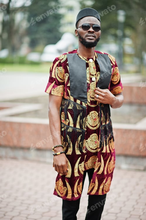 Preview: African stylish and handsome man in traditional outfit and black cap standing outdoor.