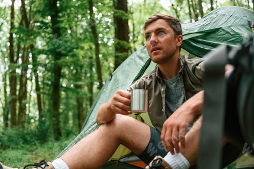 Preview: With cup of water. Sitting near the tent. Tourist in summer forest. Conception of exploration