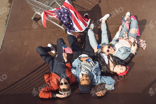 Preview: elevated view of teenagers having fun and lying with american flag in skateboard park, hipster style