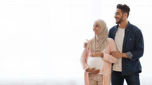 Preview: Young Pregnant Arabic Family Standing Near Window At Home And Looking Aside