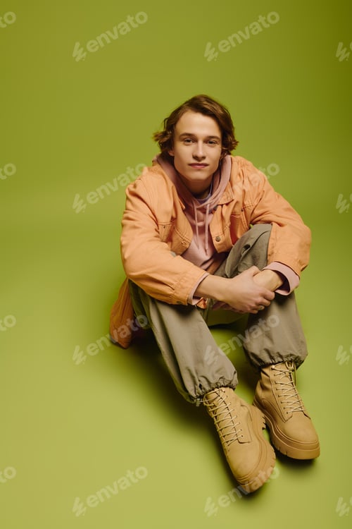 Preview: Fashionable young man sits confidently on vibrant green backdrop showcasing modern youth style