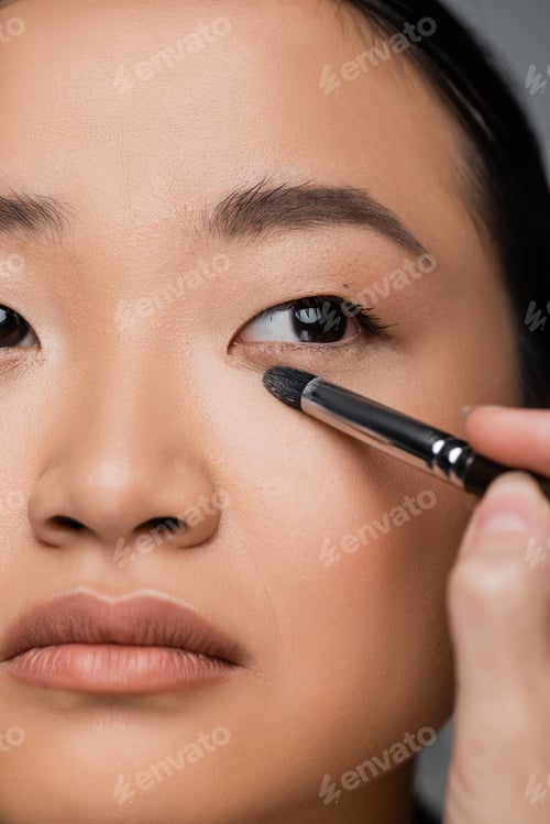 Preview: close up view of cropped asian woman with makeup foundation applying eye shadow isolated on grey