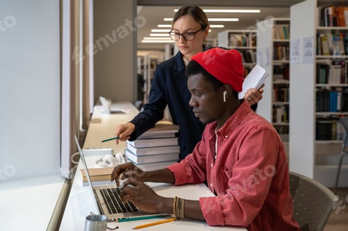 Preview: Woman teacher pointing at laptop screen while explaining something to African student guy in library