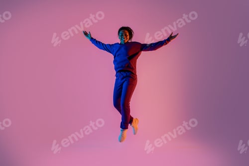 Preview: Excited black woman dancing and jumping over pink background in neon light, looking and smiling at