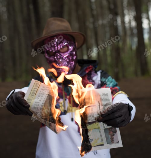 Preview: Unknown person wearing a scary face mask burning the newspaper in a forest - crime concept