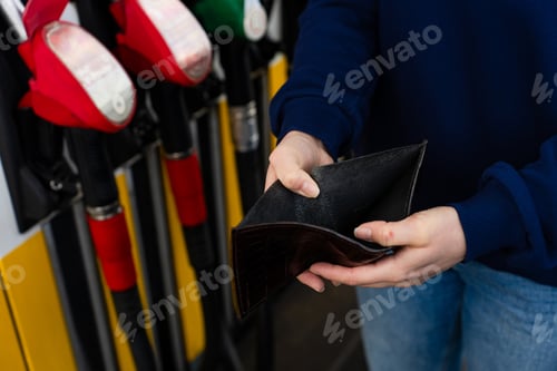 Preview: Hand holding empty wallet and gasoline nozzle at fuel station.