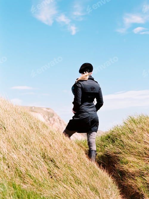 Preview: A woman in a coat and beret standing among the sand dunes by the coast.