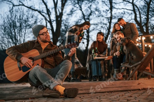 Preview: Man with guitar is sitting in front of his friends. Group of people is spending time together