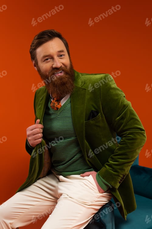 Preview: Smiling bearded man in stylish outfit sitting on armchair on red background