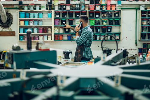 Preview: Male worker mixing colors for screen printing in a workshop and using a smartphone