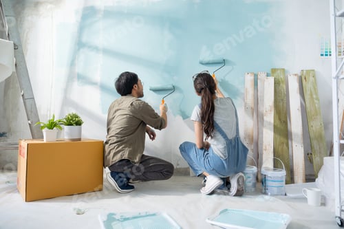 Preview: Asian couple repairing and painting the wall with blue paint using a roller during renovation